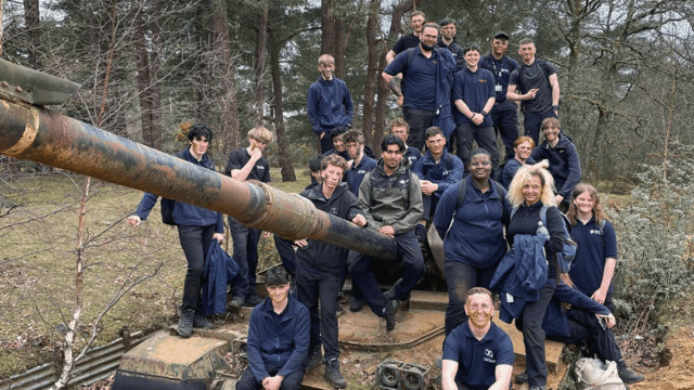 A group of young people and instructors in navy uniforms posing on and around a derelict tank in a woodland setting