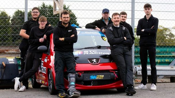 Seven members of BTC Racing team posing around a red Peugeot race car at a circuit