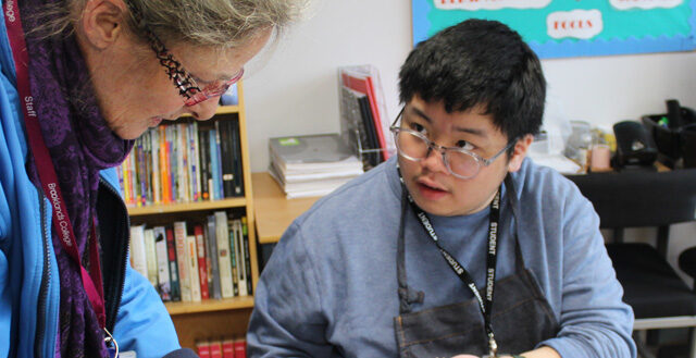 A staff member leaning over to assist a student wearing glasses with their work at a desk in a classroom