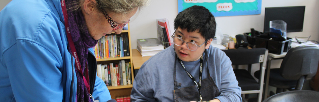 A staff member leaning over to assist a student wearing glasses with their work at a desk in a classroom