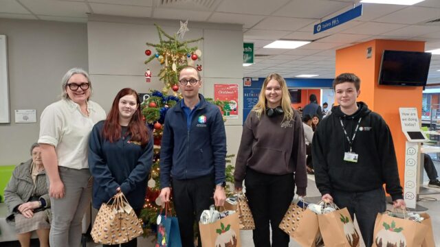 Five people holding Christmas gift bags pose in front of a decorated Christmas tree in a public building