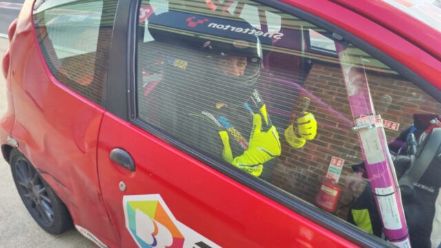 A helmeted racing driver in a yellow race suit gives a thumbs up from inside a red race car at Snetterton circuit