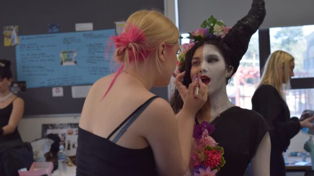 A makeup artist applies lipstick to a model in Maleficent-inspired costume with large black horns and white face paint