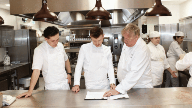Three chefs in white uniforms reviewing documents at a stainless steel kitchen counter in a professional restaurant kitchen