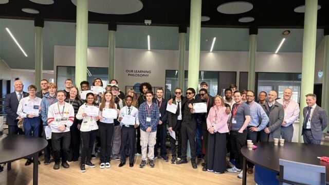 A large group of students and staff posing together indoors, several students holding certificates, in front of an "Our Learning Philosophy" sign