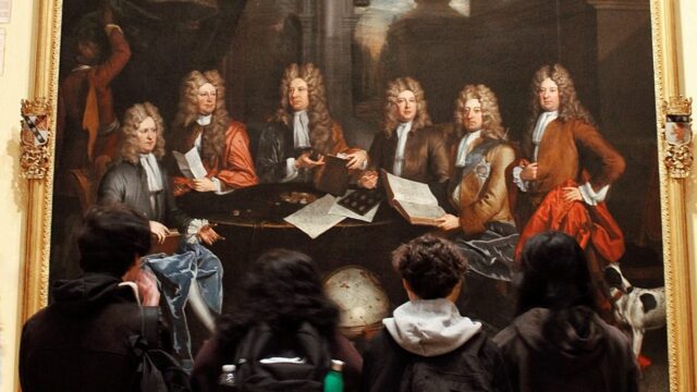 Students sitting on the floor studying a large ornate oil painting depicting a group of 17th-century gentlemen in wigs gathered around a table