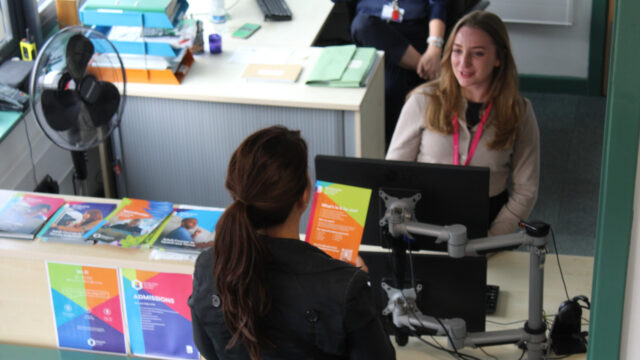 A staff member wearing a lanyard assists a visitor at a college admissions reception desk, with leaflets and an Admissions poster visible
