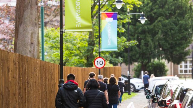 People walking along a tree-lined street with colourful lamp post banners reading "Collaborative" and "A Champion