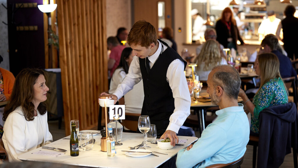 A waiter serves a couple at table 10 in a busy restaurant dining room with an open kitchen visible in the background
