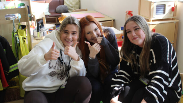 Three smiling students wearing lanyards pose together in a nursery or early years classroom setting