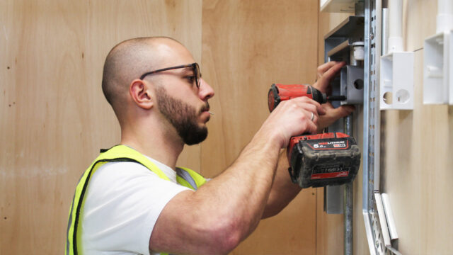 A tradesperson in a hi-vis vest using a cordless drill to fit an electrical back box to a wall-mounted metal frame
