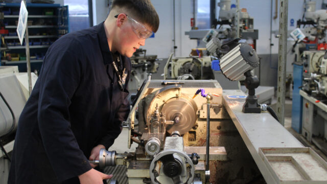 A young apprentice wearing safety goggles and a navy boiler suit operates a metal lathe in a workshop