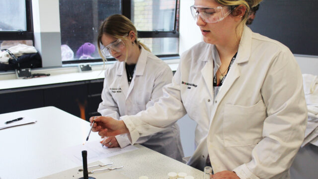 Two Brooklands College students in lab coats and safety goggles conducting a science experiment in a laboratory