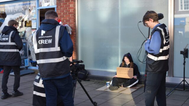 Futureframe Productions crew filming a young person sitting on the pavement holding a cardboard sign, outside a shop on a high street