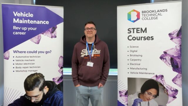 A smiling man wearing a lanyard stands between two Brooklands Technical College banners promoting Vehicle Maintenance and STEM Courses