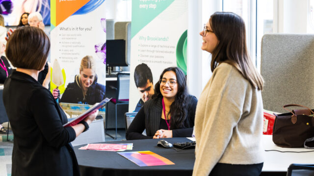 A smiling Brooklands College adviser at an information stand speaks with visitors, with apprenticeships and transition banners displayed behind her