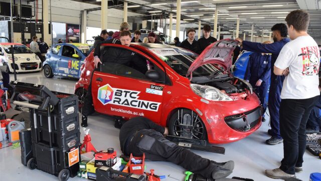 Students working on a BTC Racing car branded with brooklands.ac.uk in a motorsport garage, with tools and other race cars visible