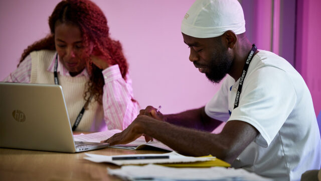 Two students wearing lanyards studying together at a desk with a laptop and printed documents