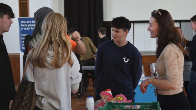 Young people chatting with an adviser at an information stand with a crate of supplies at what appears to be a community event or fair