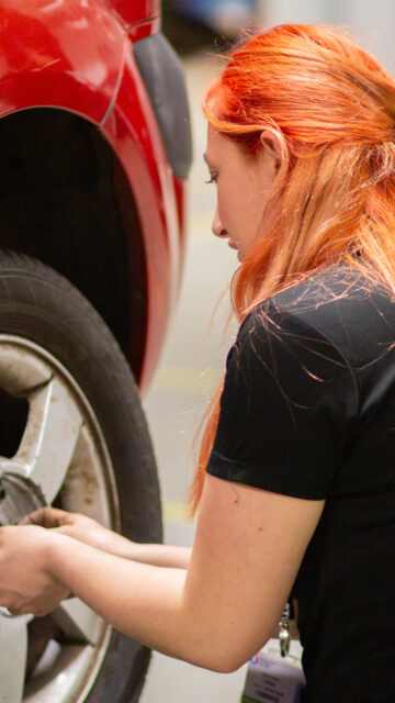 A young woman with red hair working on the wheel of a red car in a garage or workshop setting