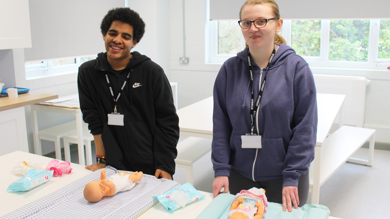 Two students wearing lanyards practising nappy changing on baby dolls in a childcare classroom
