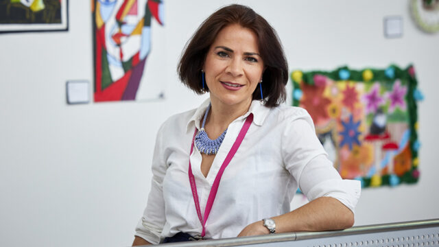 Woman wearing a white shirt and lanyard, smiling in a school or college setting with colourful artwork displayed on the wall behind her