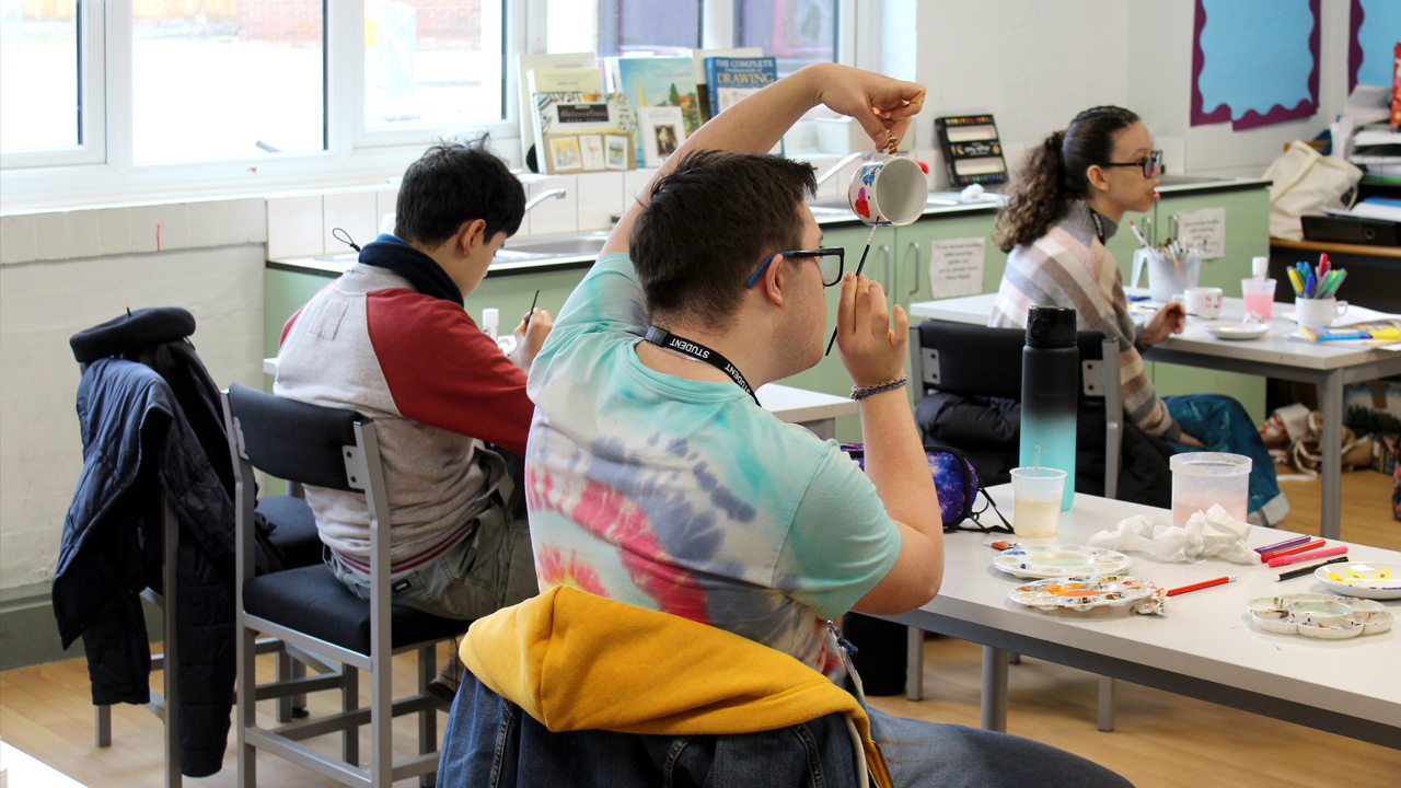Students painting mugs in an art classroom, with paint palettes, brushes, and art supplies on the tables