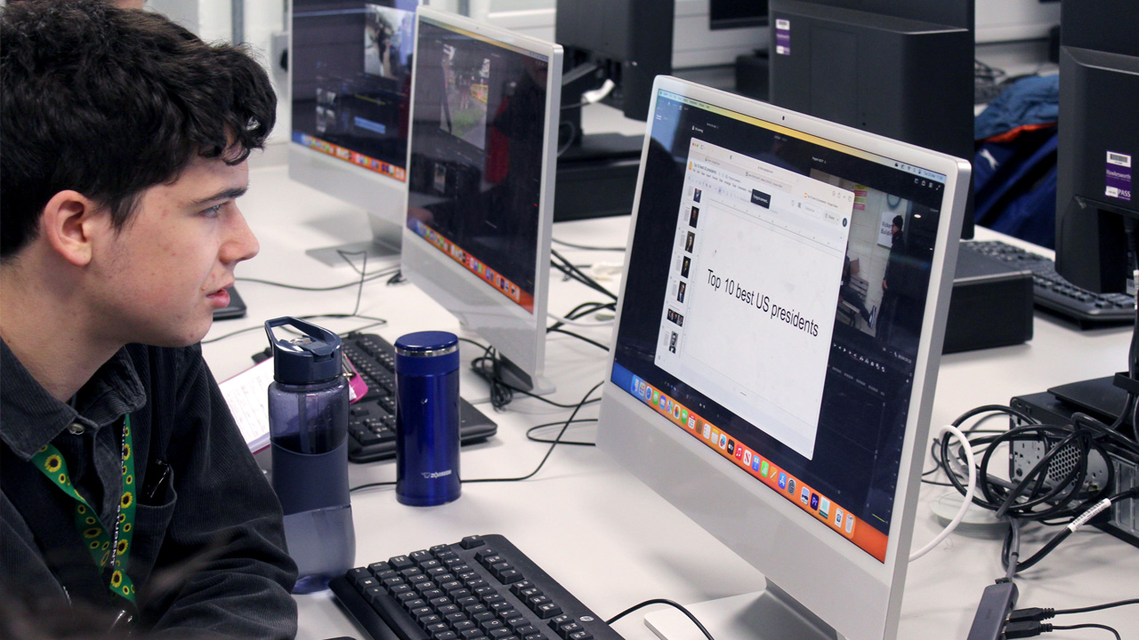 Student working at a Mac computer in a classroom, with a document titled "Top 10 best US presidents" on screen