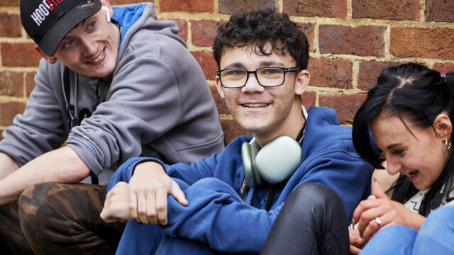 Three college students sitting together against a brick wall, smiling and relaxed, wearing lanyards and headphones