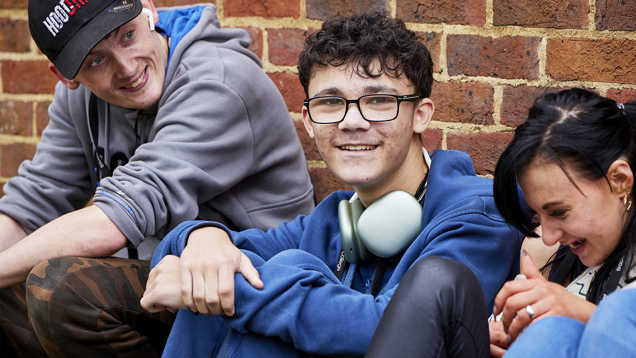 Three college students sitting together against a brick wall, smiling and relaxed, wearing lanyards and headphones