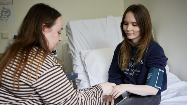 A healthcare professional taking a patient's blood pressure in a hospital room, with the patient seated on a bed
