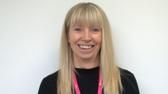 A smiling woman with long blonde hair wearing a black top and a pink Brook staff lanyard