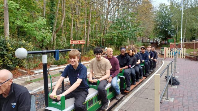 A group of young people and adults riding a miniature railway train at Twichen Manor station, surrounded by autumn trees