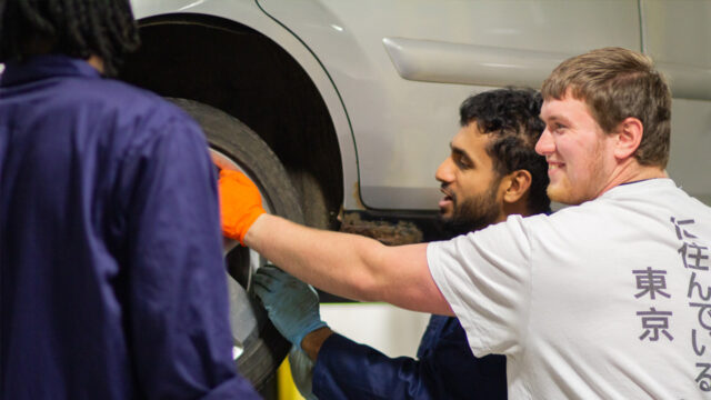 Three people working together on a vehicle tyre in a garage, with two wearing protective gloves