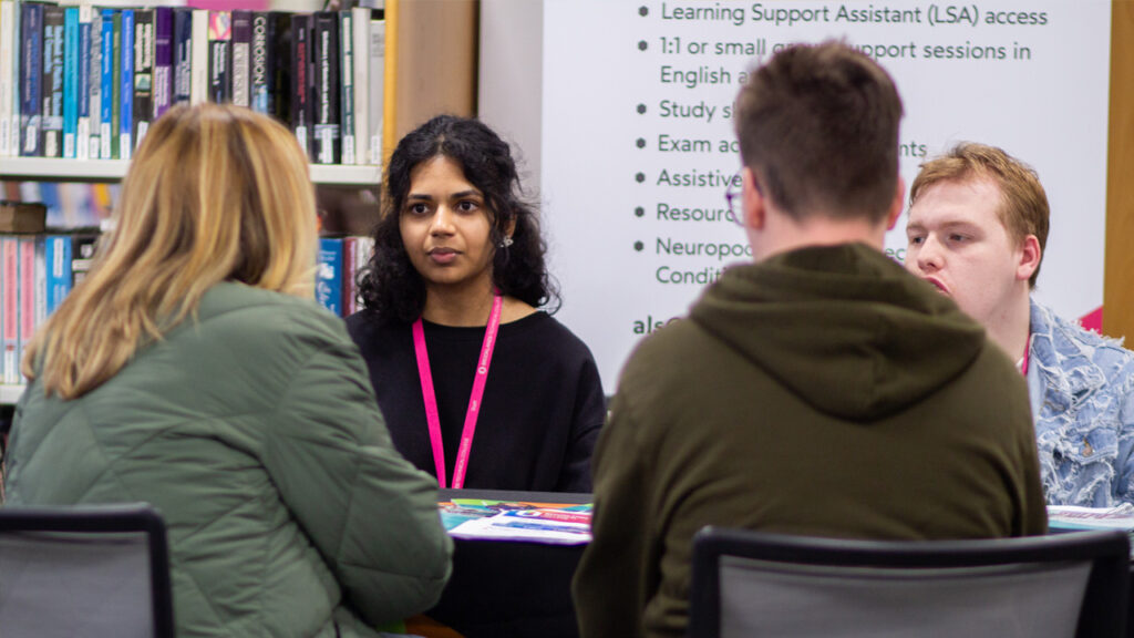 A college staff member wearing a lanyard speaks with students at a table, next to a banner listing learning support services