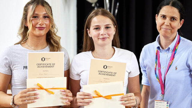 Two students holding Brooklands Technical College Certificates of Achievement, presented by a member of staff wearing a college lanyard