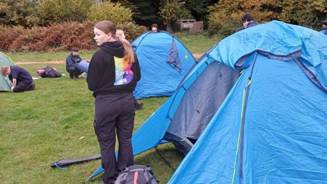 Young people setting up blue dome tents on a grassy campsite surrounded by autumn trees