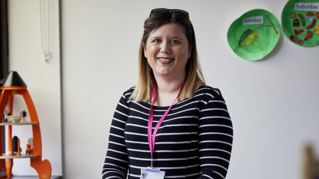 A smiling woman wearing a striped top and pink lanyard with ID badge, standing in a primary school classroom