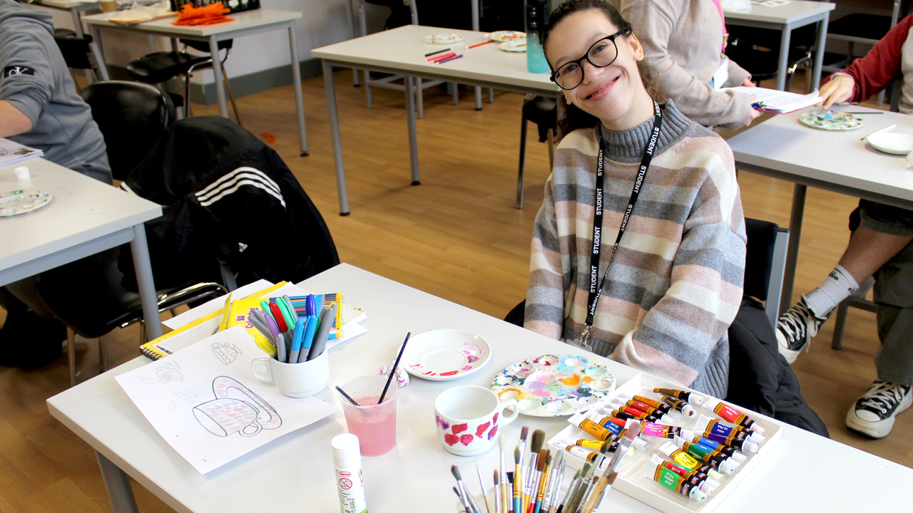 A smiling student wearing glasses and a lanyard sits at an art class table with paints, brushes, palettes, and a sketch in progress