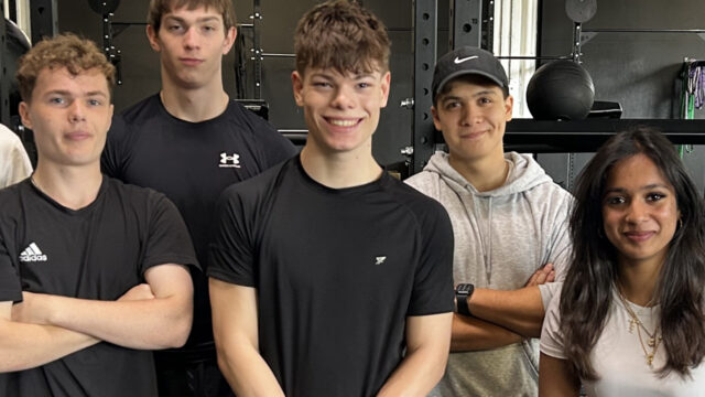 Five teenagers posing together in a gym, wearing sportswear, with weight training equipment visible in the background