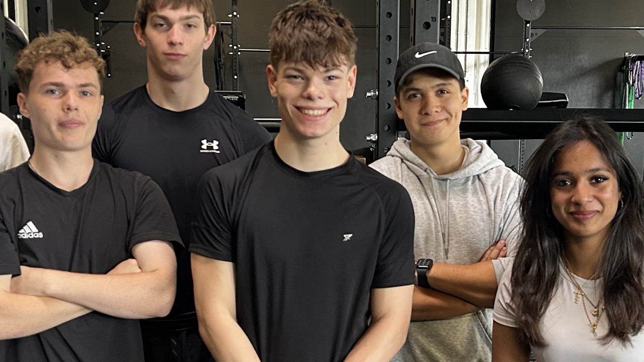 Five teenagers posing together in a gym, wearing sportswear, with weight training equipment visible in the background