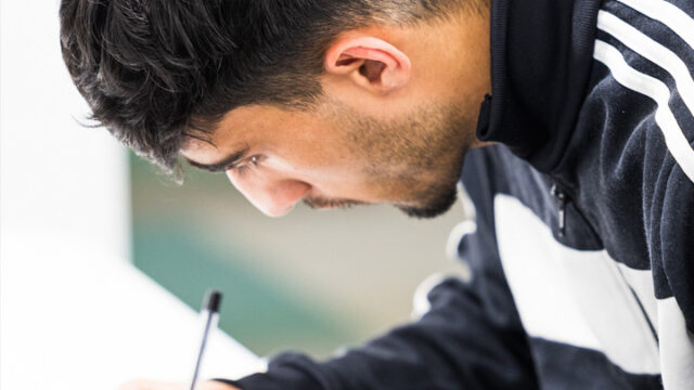 Young man wearing glasses and a sports jacket, leaning over a desk and writing with a pen
