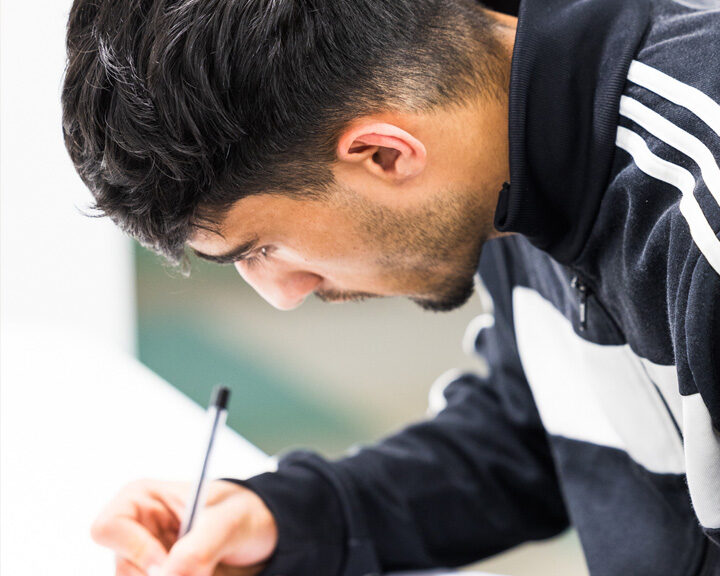 Young man wearing glasses and a sports jacket, leaning over a desk and writing with a pen