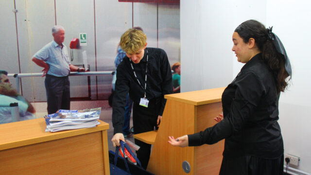 Two students wearing lanyards handling leaflets and a tote bag at a reception desk in a college or university setting