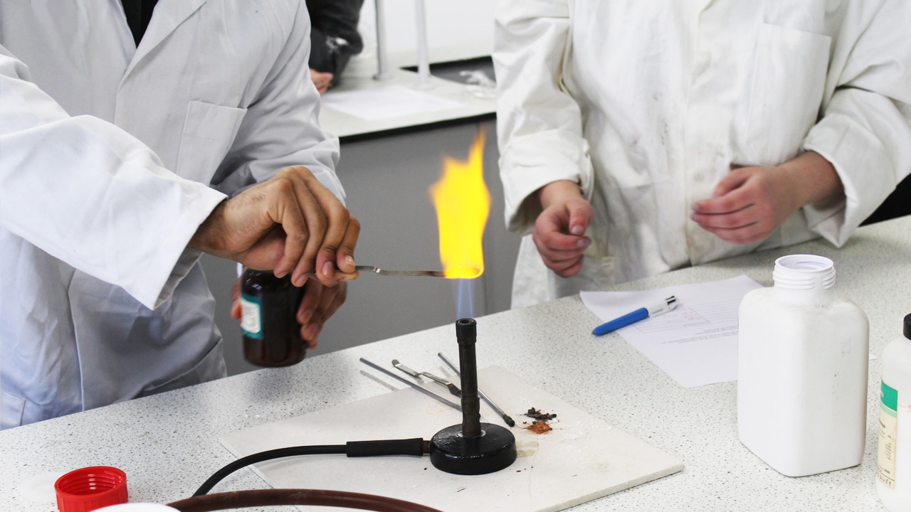 Students in lab coats conducting a flame test experiment with a Bunsen burner in a science laboratory